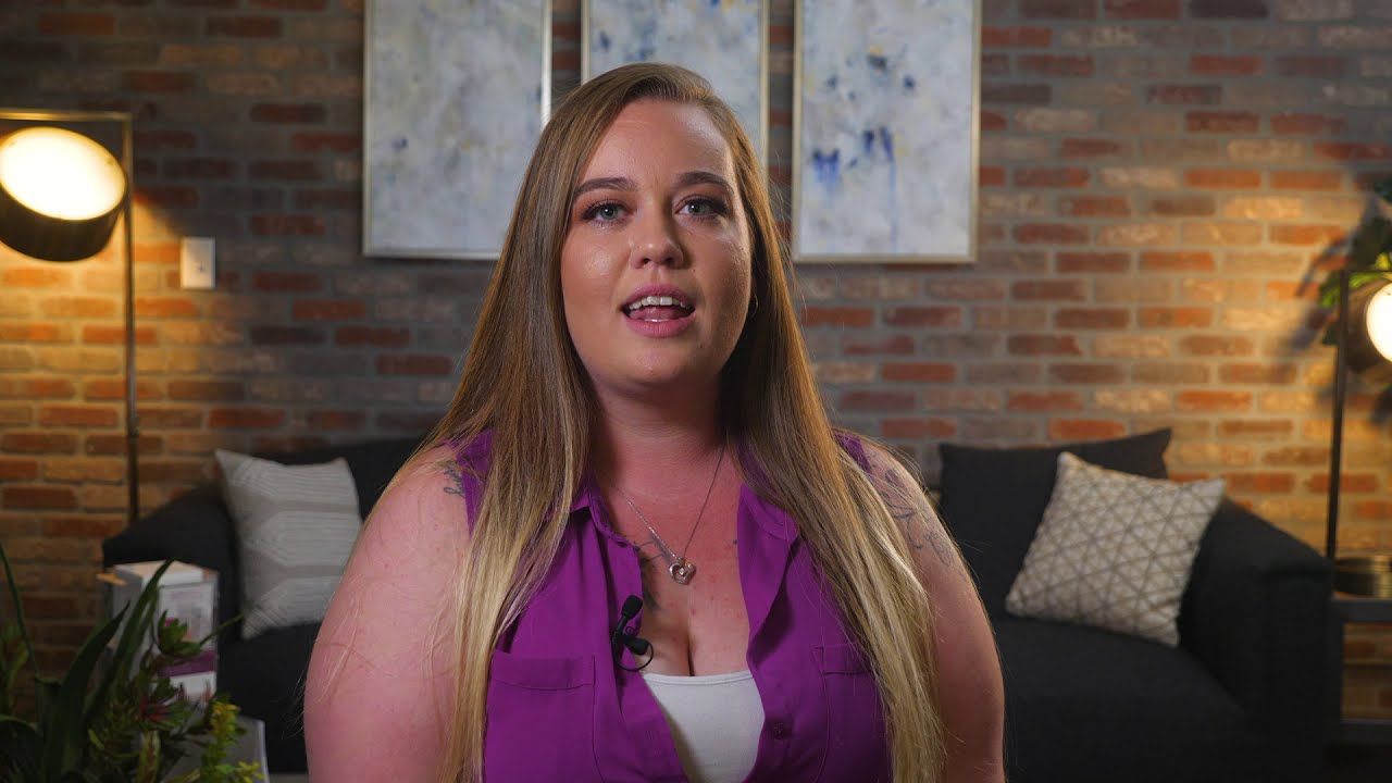 Casey, a Vein Envy patient, mid-interview on a couch in front of a brick wall, lit by two warm lamps.