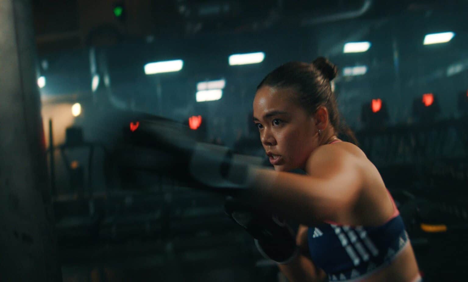 A woman in boxing gloves and an adidas sports top trains in a dark gym lit by strip lights.