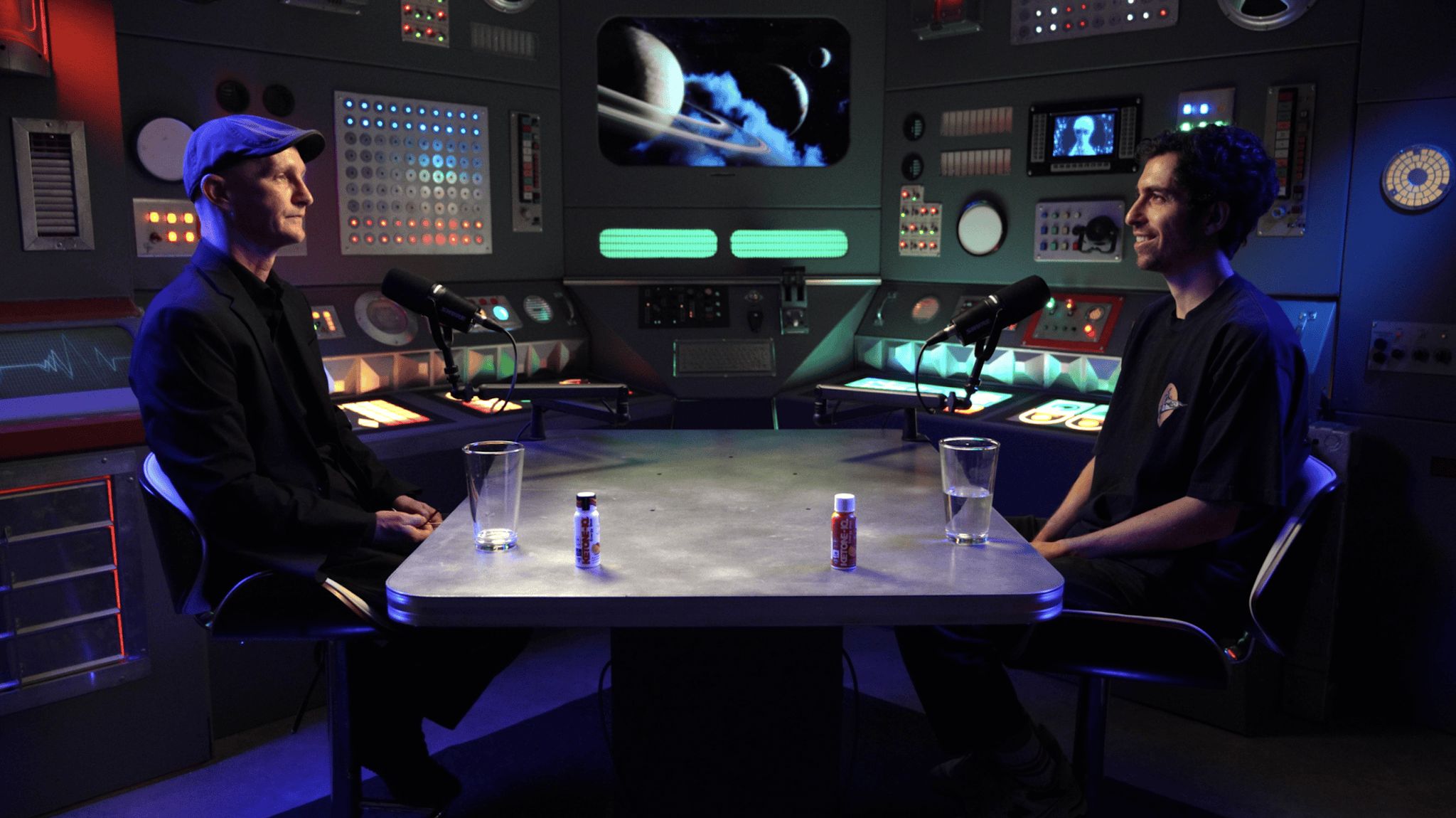 Two hosts face each other across a steel table on the American Alchemy set, lit by blue and teal control-room panels.