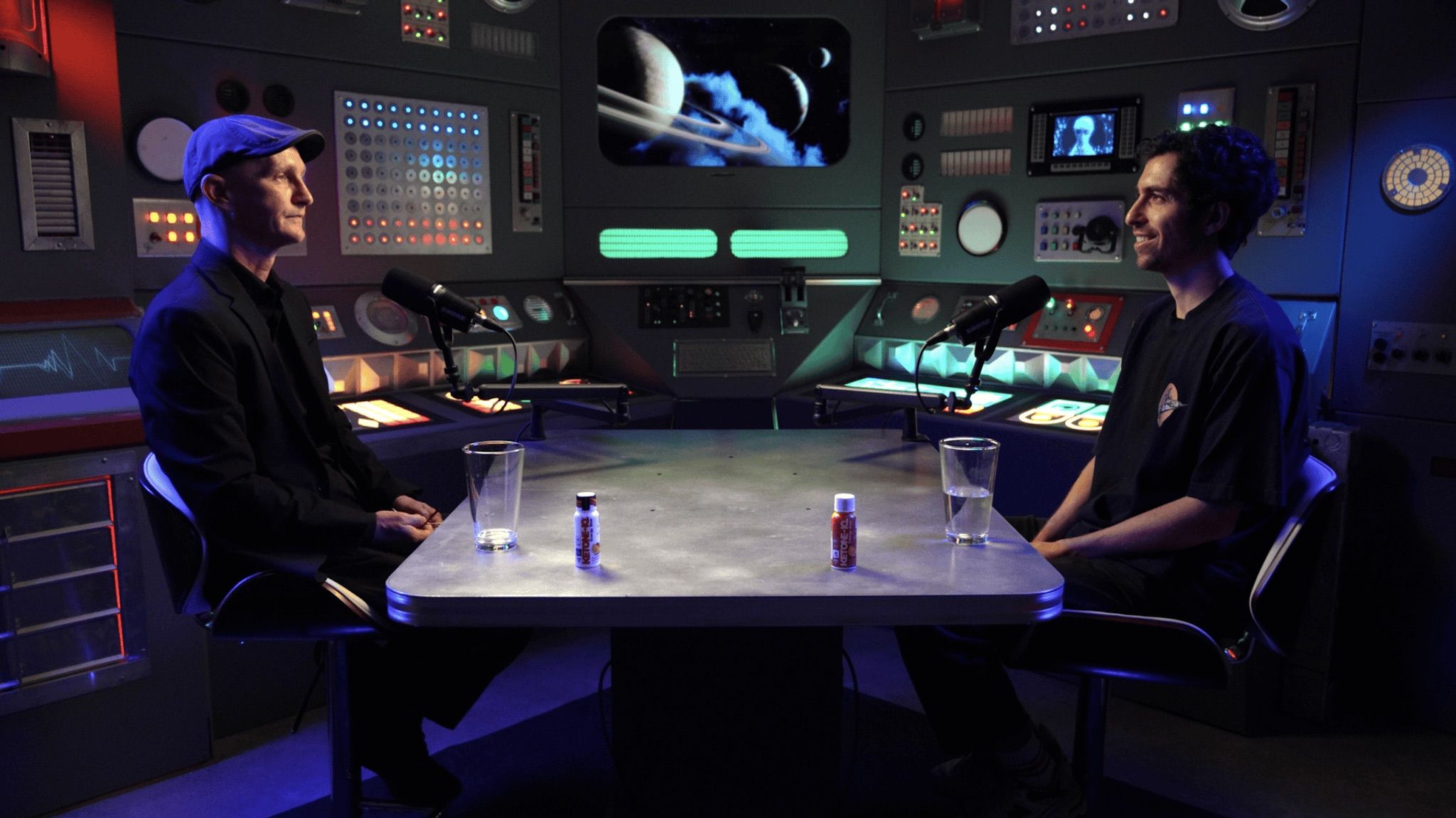 The finished American Alchemy podcast set: two hosts at the table in front of the lit console wall