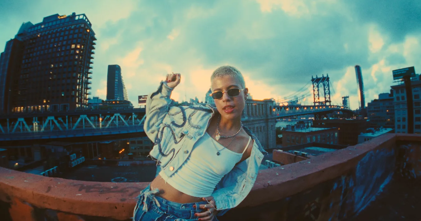 A woman in sunglasses and a white crop top dances on a rooftop, the New York skyline and a bridge behind her.