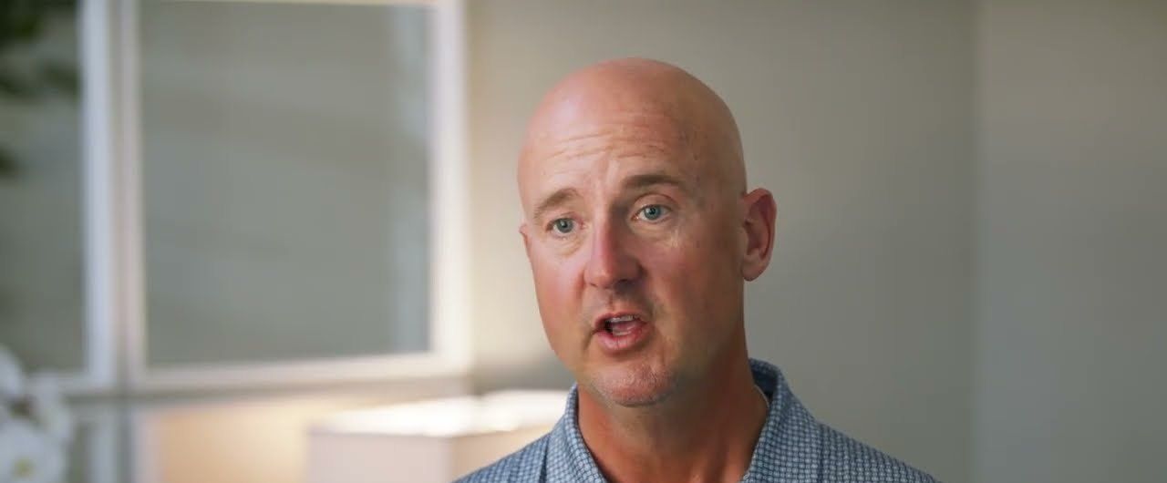 Chad, a bald man in a patterned blue shirt, talks during his interview in a softly lit room.