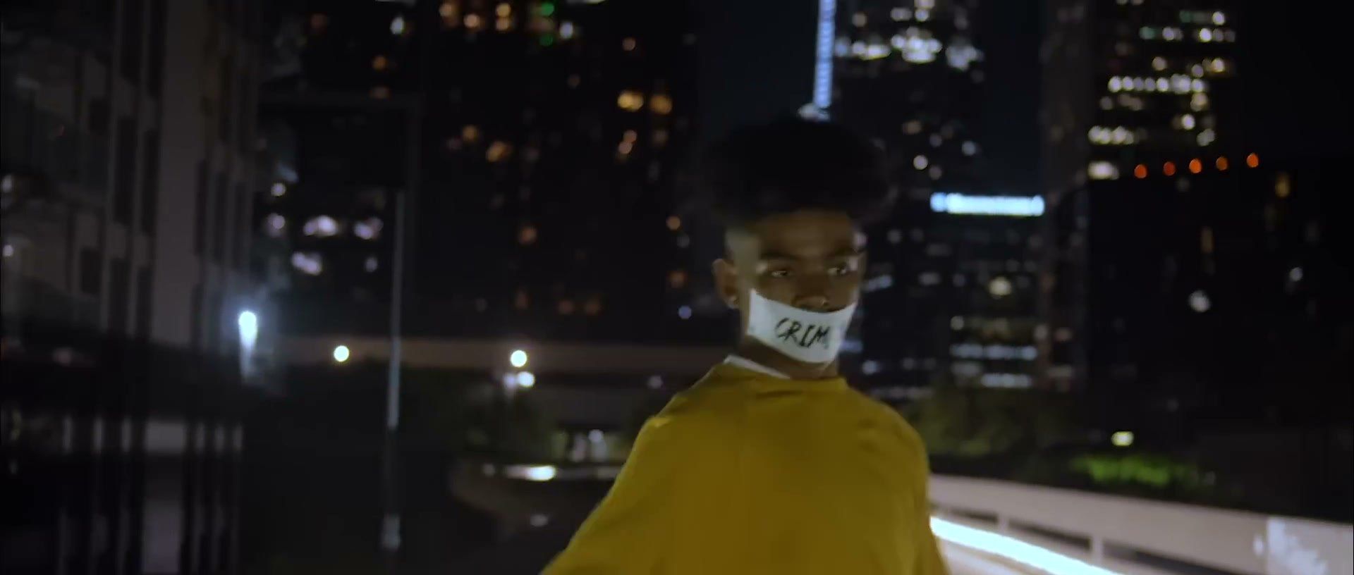 A person in a yellow jacket and white face mask stands on a rooftop above a city skyline at night.
