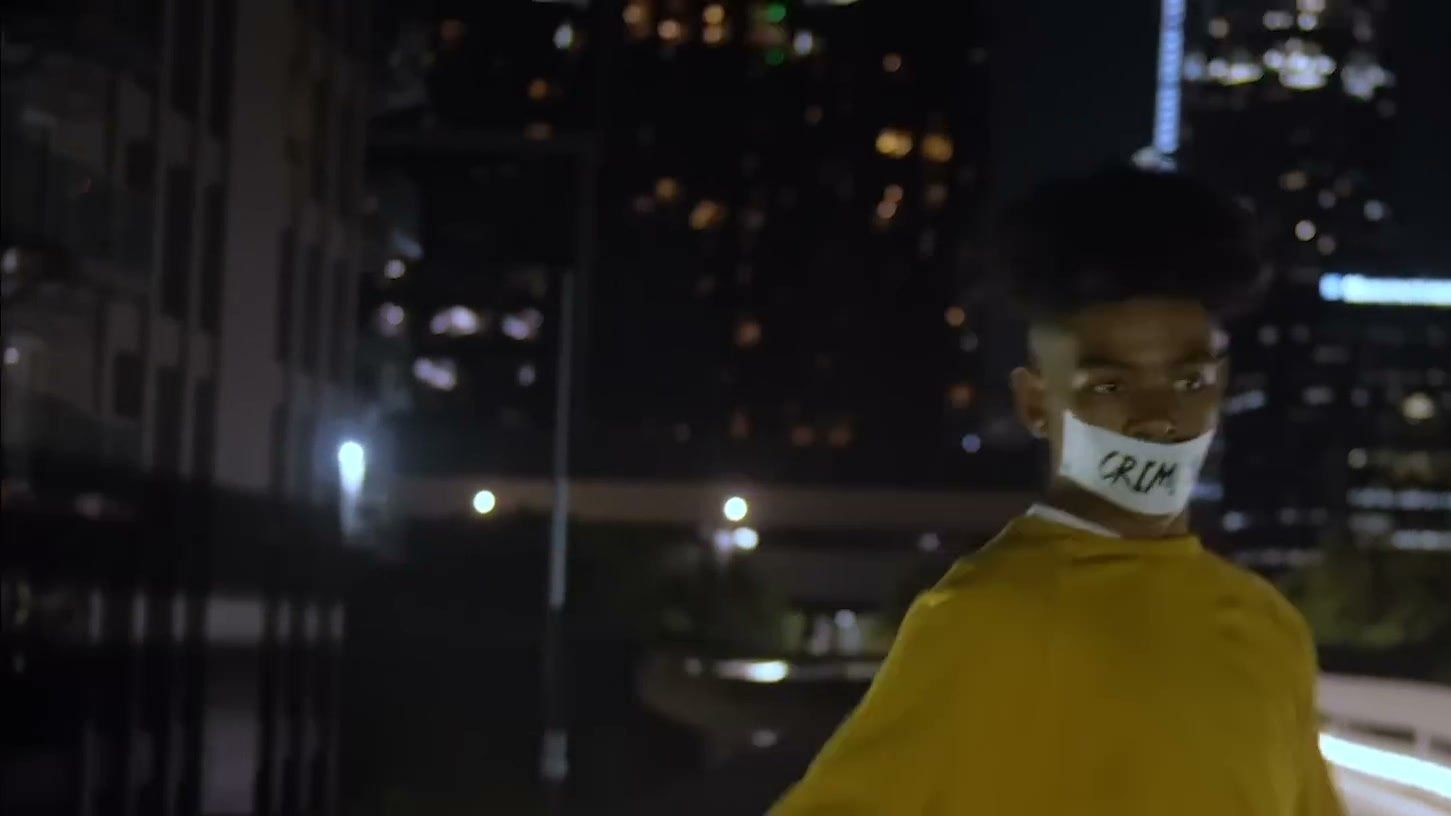 A person in a yellow jacket and a white face mask stands on a rooftop above the downtown Los Angeles skyline at night.