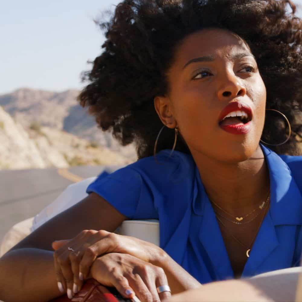 A singer with curly hair and red lipstick rides in an open car through the desert, a still from CHERONE’s “Lavender Sky”.