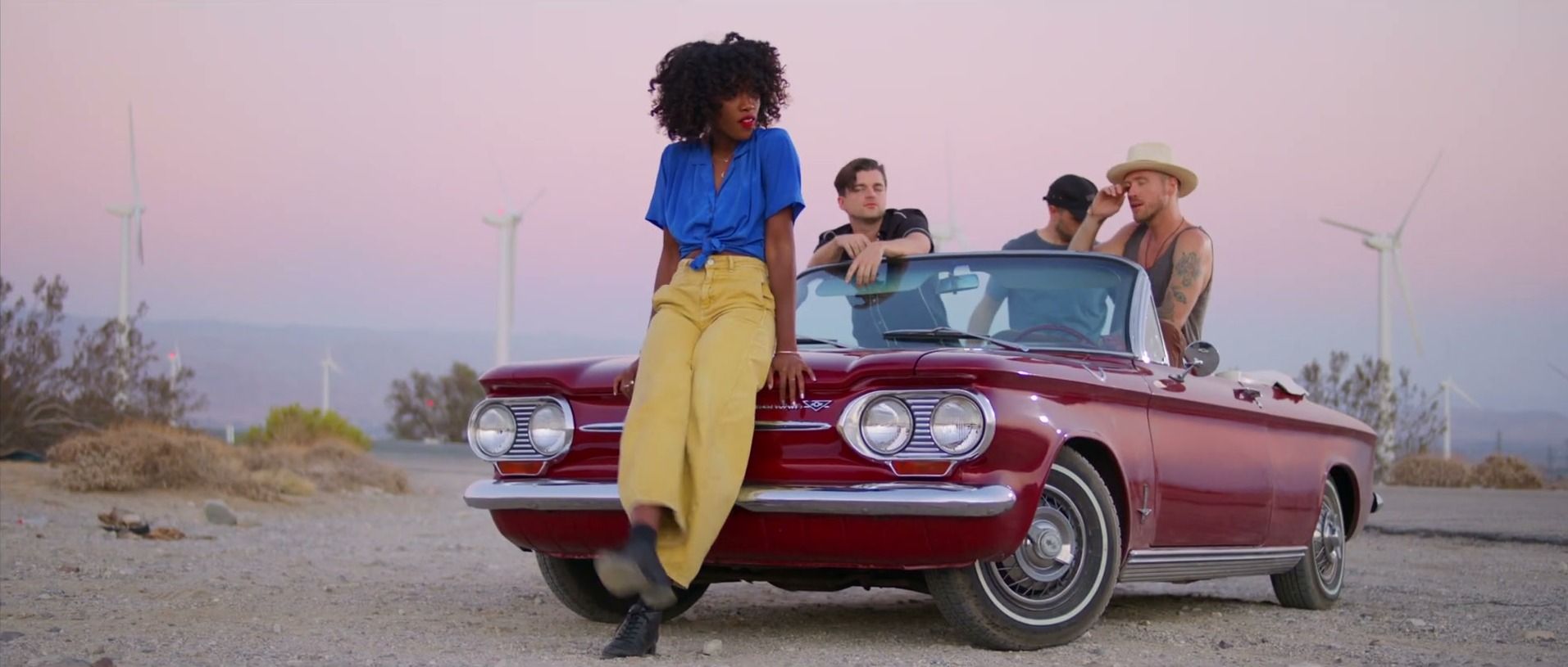 A singer in a blue shirt and yellow trousers leans on a red convertible at dusk, her band in the car behind her and wind turbines on the hills.