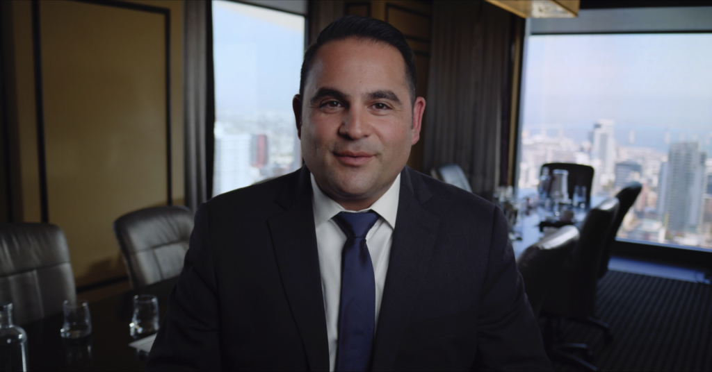 A man in a suit and tie speaks to camera in a high-rise office overlooking the city.