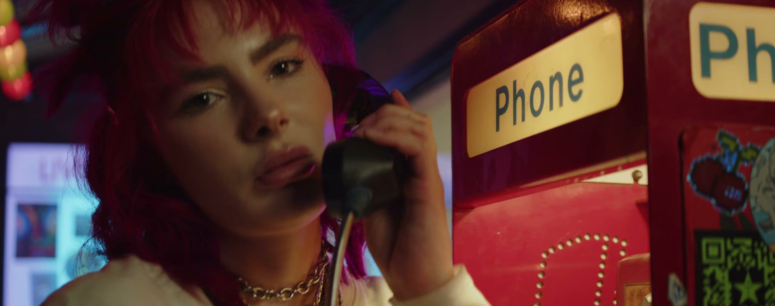 A singer with pink hair holds a payphone receiver beside a red phone booth, a still from Courtney Paige Nelson’s “CYANIDE JAWBREAKER”.