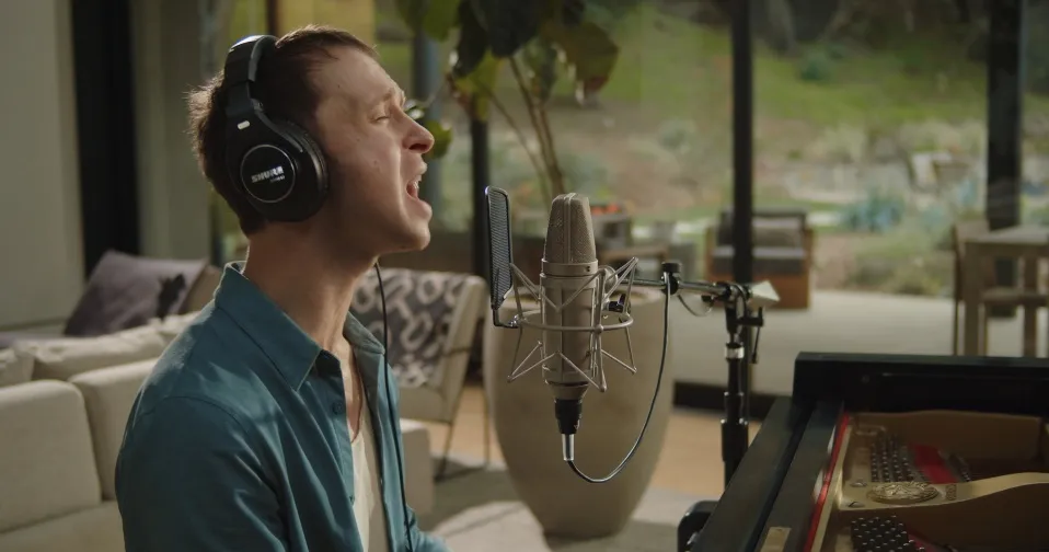 Darcy Callus sings into a studio microphone in headphones beside a grand piano, in a glass-walled house above the hills.
