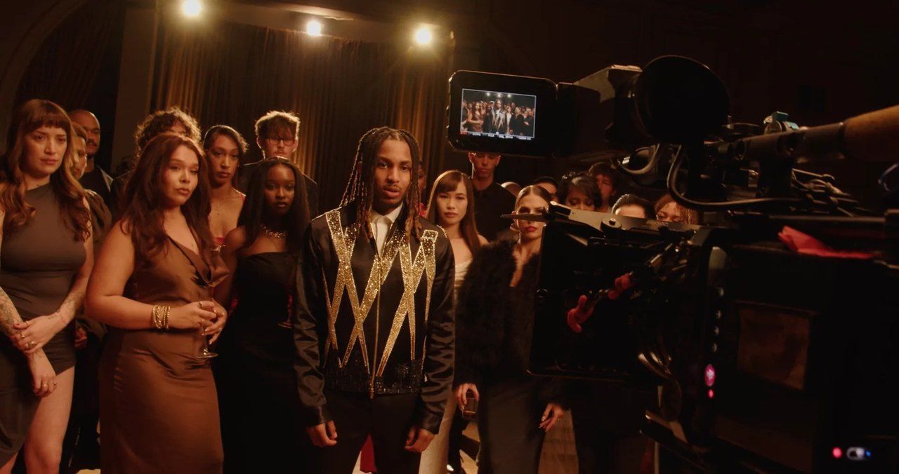 DDG stands in front of a crowd on a dark set, a cinema camera and its monitor in the foreground.