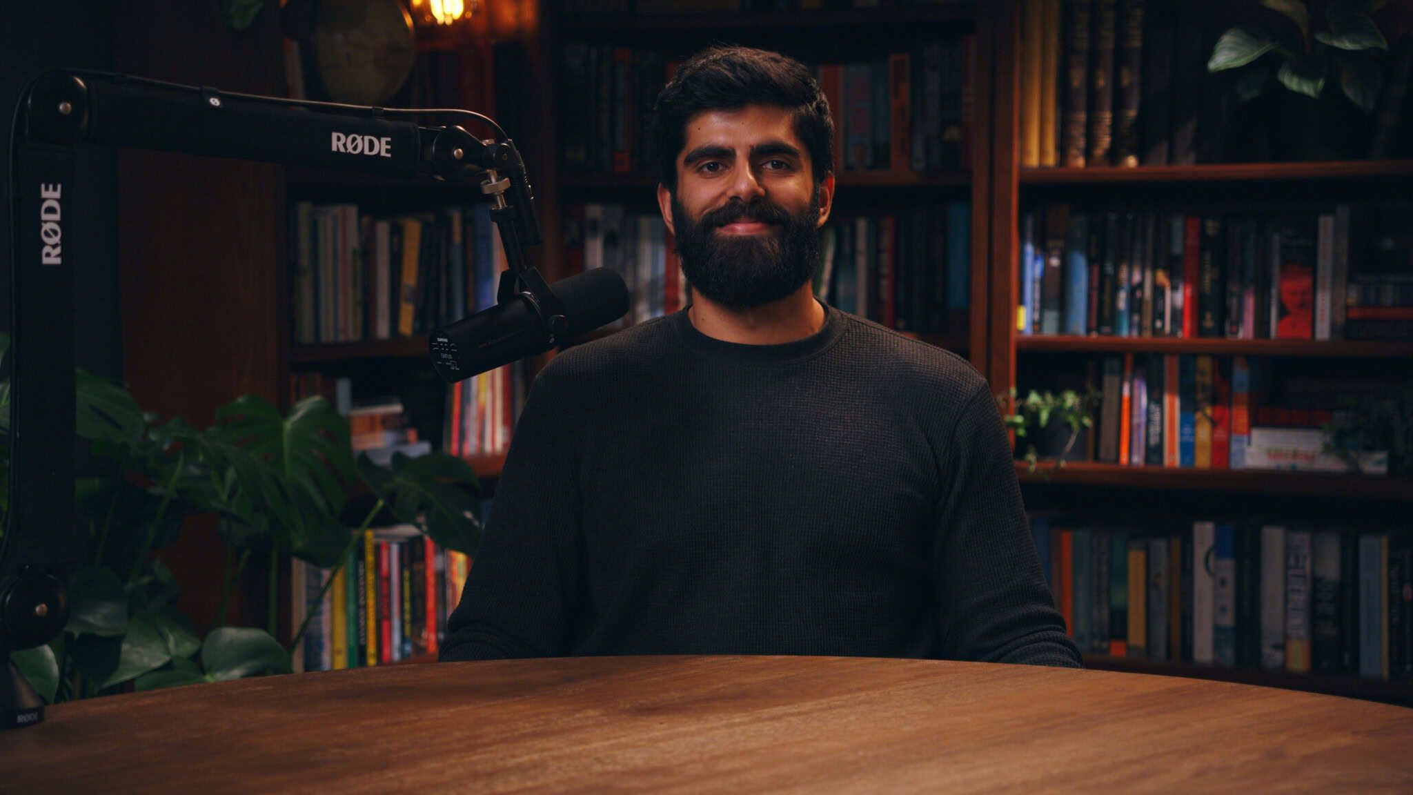 Dwarkesh Patel at his microphone in front of warmly lit bookshelves.
