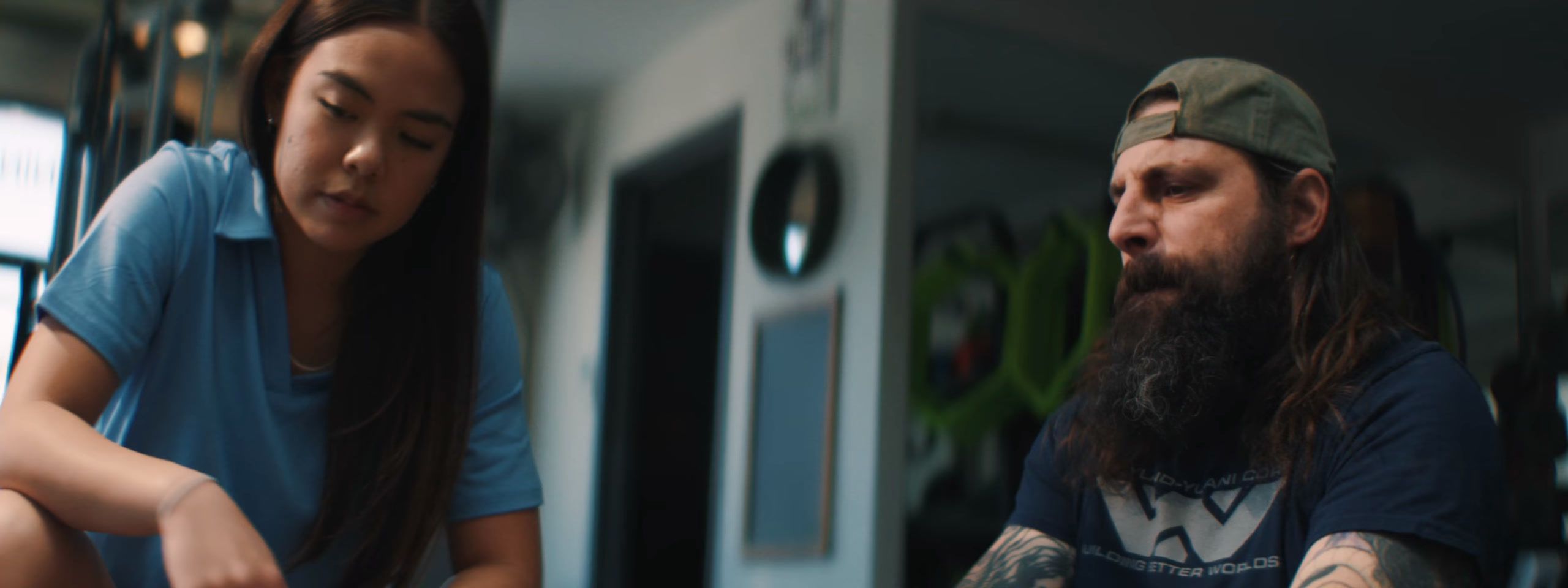 A young woman in a blue polo crouches and talks with a bearded man in a cap at the gym.