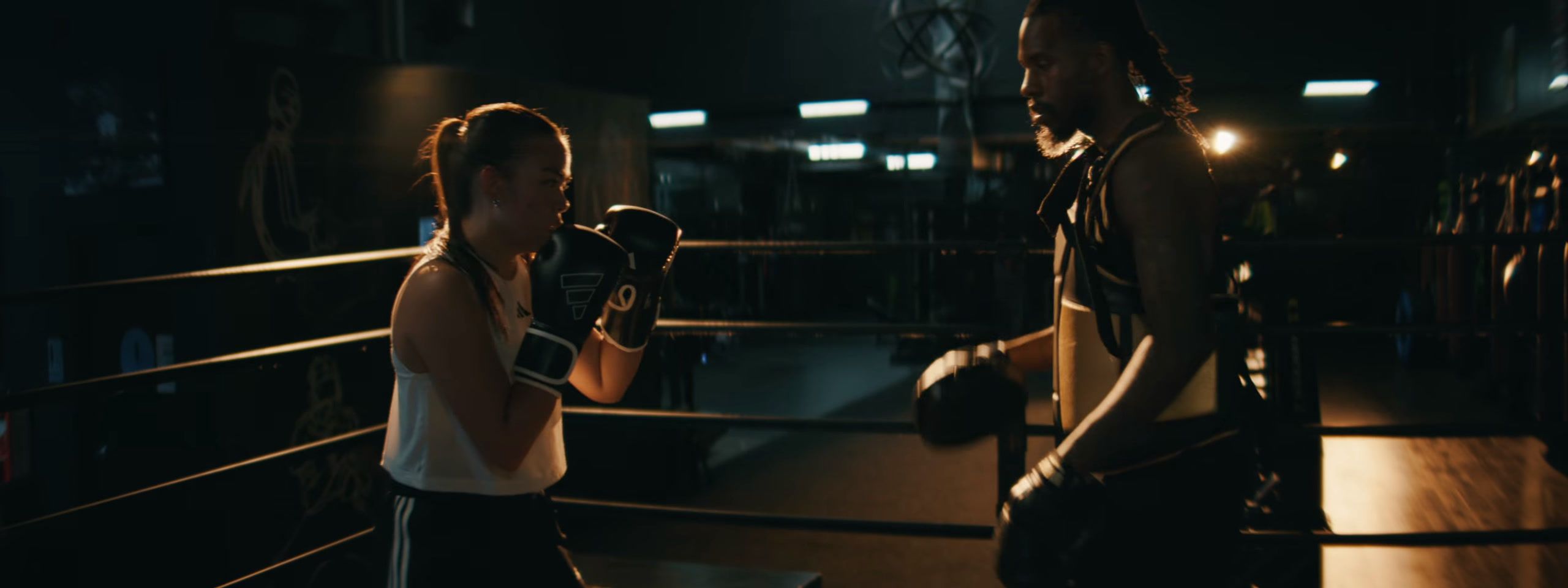 A boxer and her trainer square up in a dark gym ring.