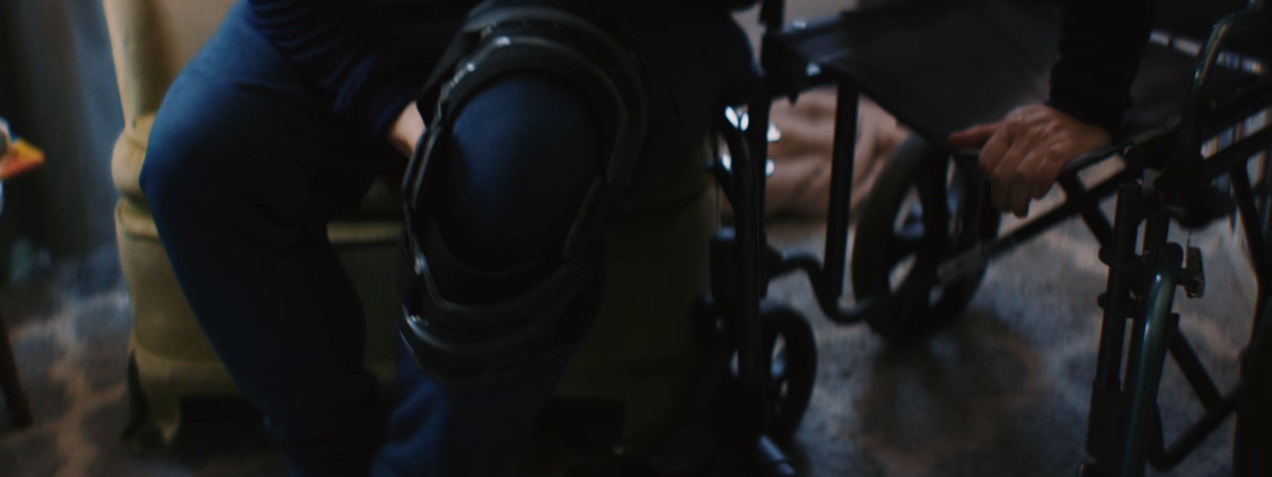 Someone in a knee brace sits beside a wheelchair, one hand on its wheel, in a dim room.