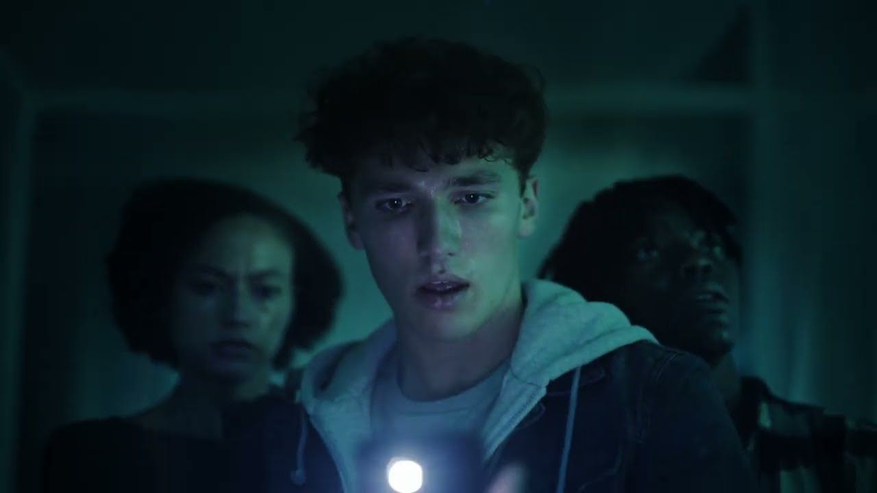Three teenagers in a dark room stare ahead in fear, lit by a flashlight.