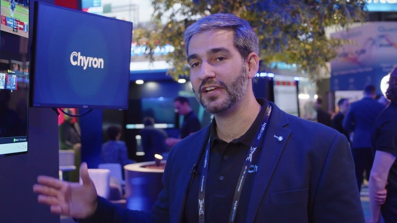 A Chyron team member talking to camera beside a screen at the company’s trade show booth