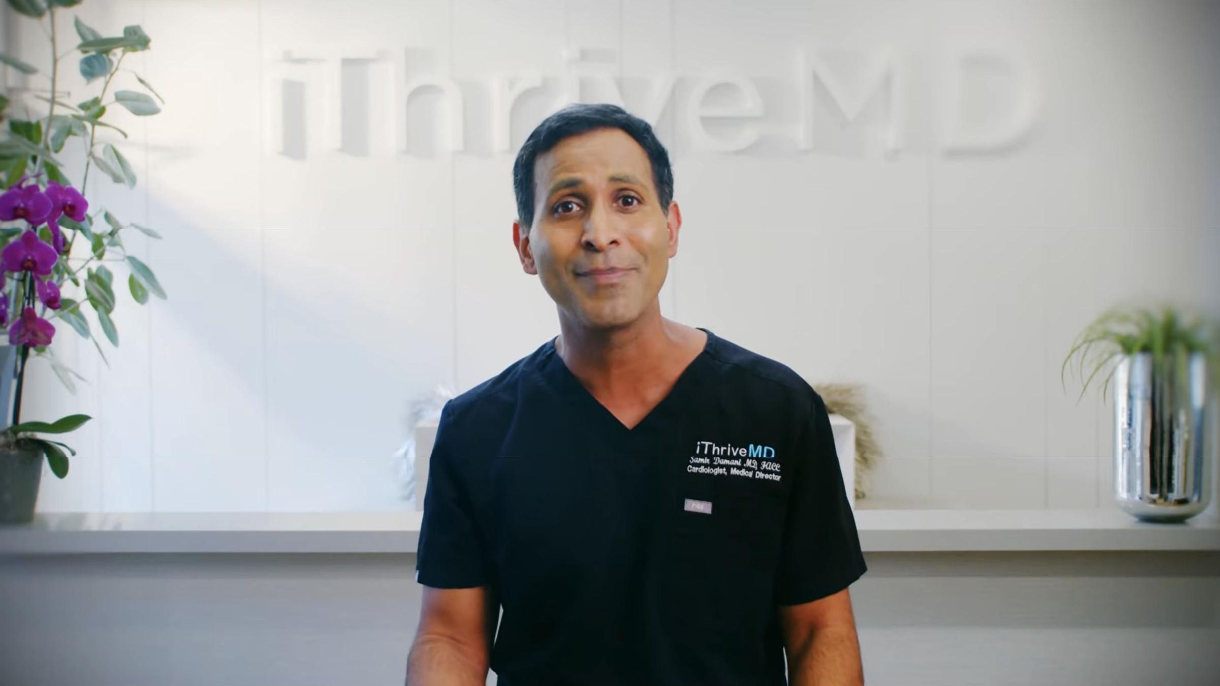 Dr. Samir Damani in dark scrubs talking to camera in front of the iThriveMD logo wall.