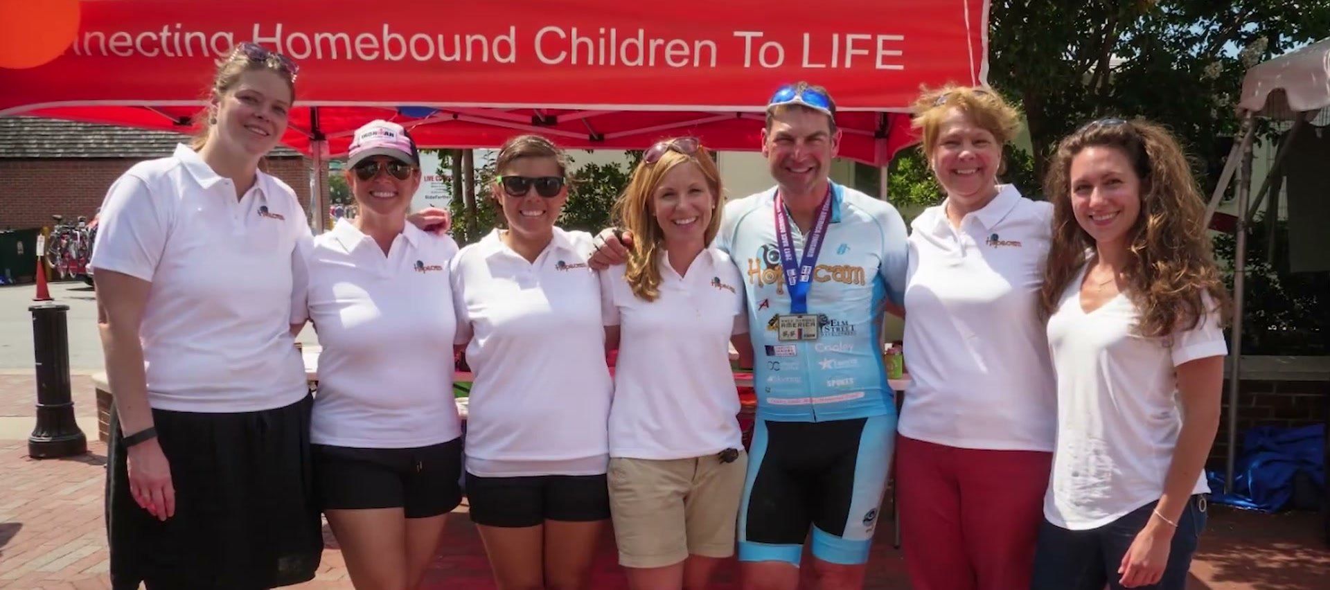 The Hopecam team in white polos stands beside a cyclist wearing a medal, under a red Hopecam tent.