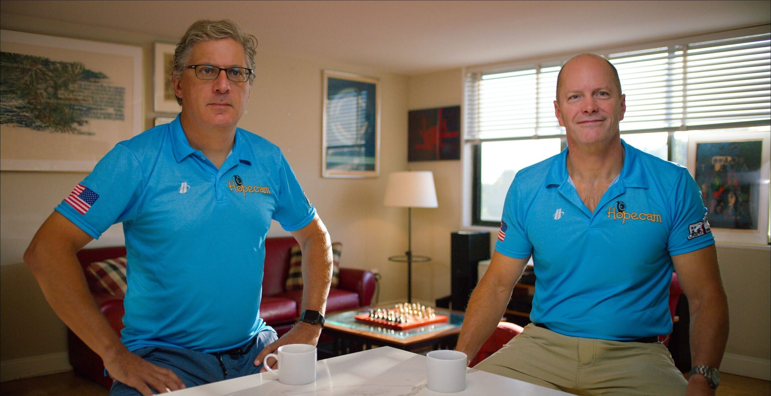 Two men in matching blue polo shirts in a living room, from Honey’s Hopecam documentary