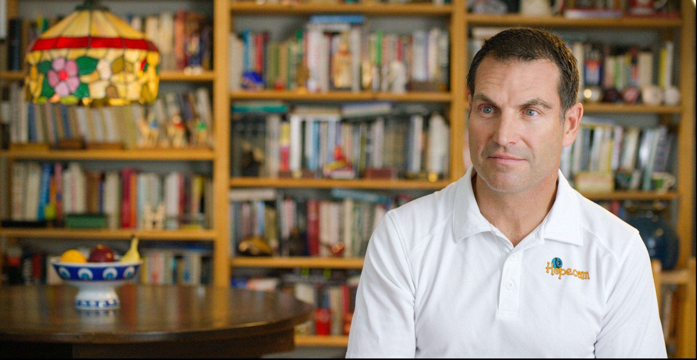 Len Forkas, Hopecam’s founder, in a white Hopecam polo, listens during his interview in front of a bookcase.