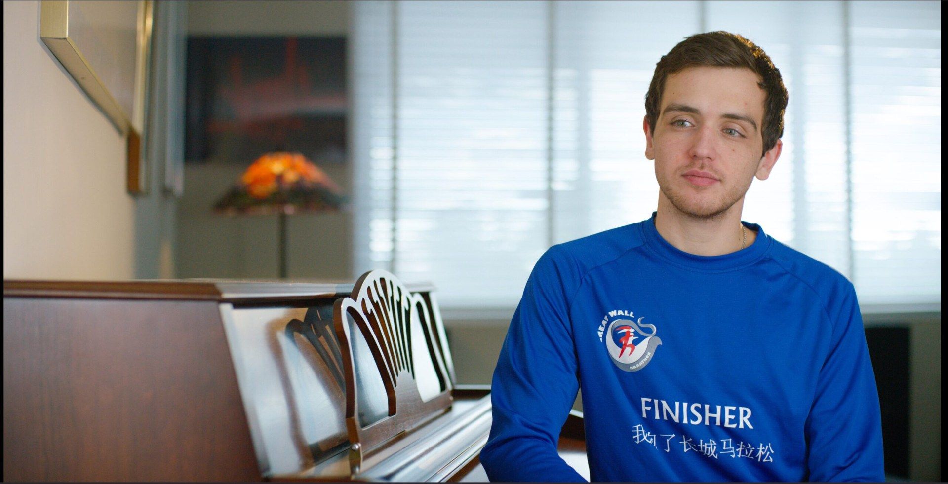 Matt, a young man in a blue marathon finisher shirt, sits by a piano during his interview.