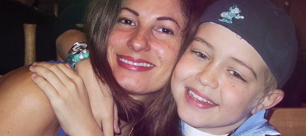 A mother hugs her smiling son, cheek to cheek, in a warm close-up.