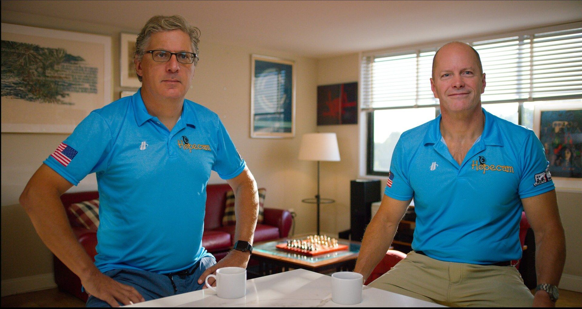 Two men in blue Hopecam polos lean on a table in a living room with a chess set behind them.