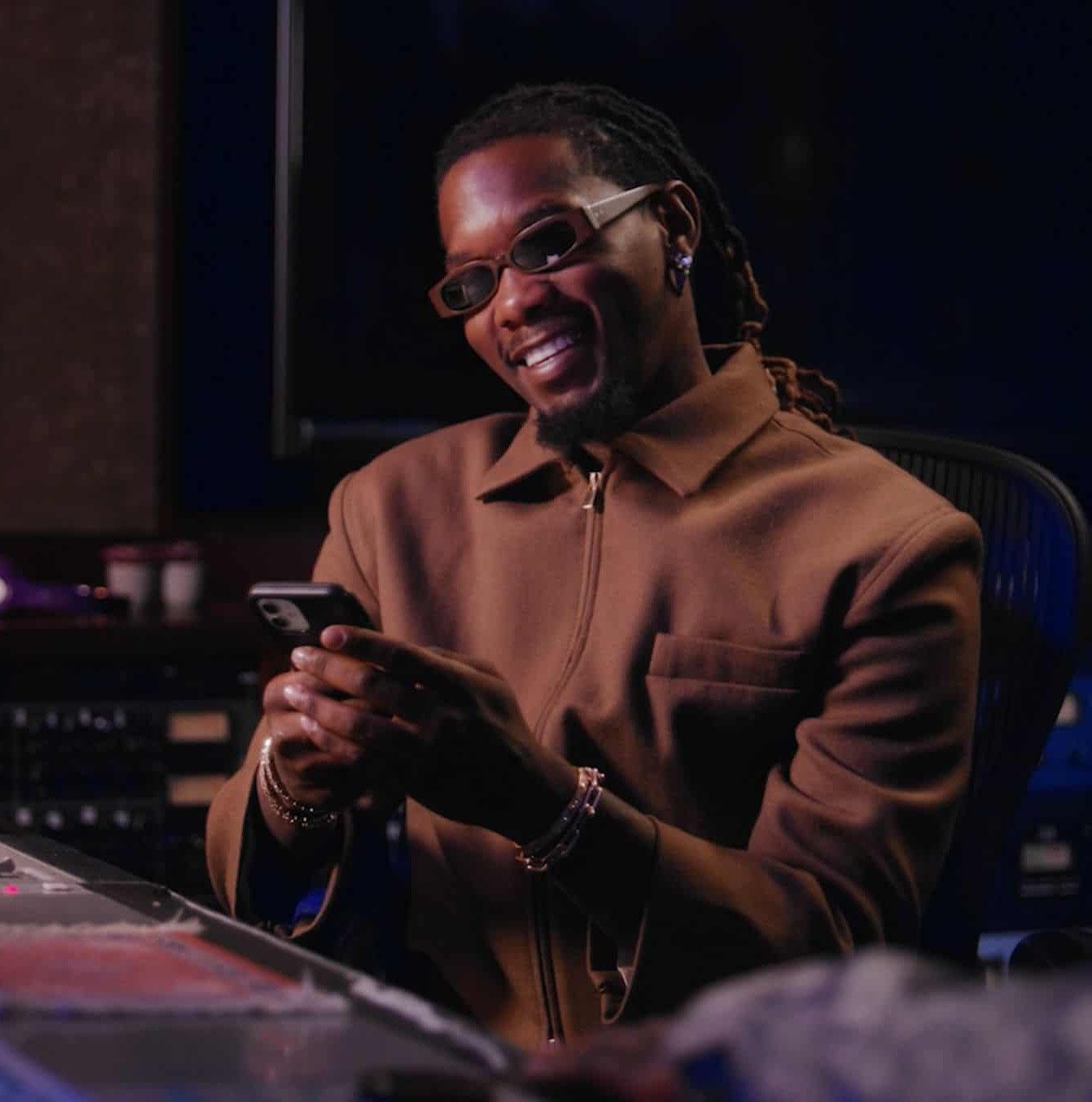 A man in tinted sunglasses and a brown jacket smiles at his phone beside a studio mixing console.