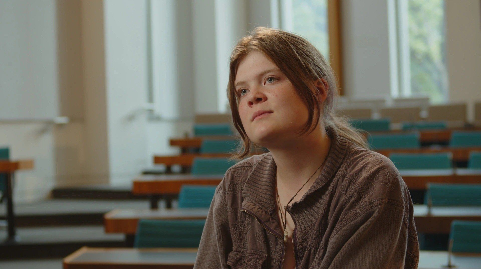 A young woman in a brown cardigan looks off camera during an interview in an empty lecture hall.