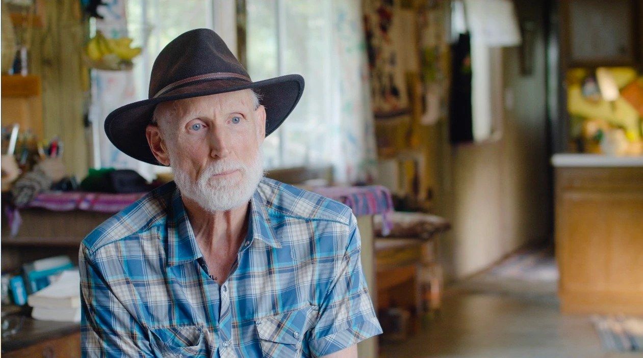 A white-bearded man in a black cowboy hat and plaid shirt sits for an interview in a sunlit workshop.