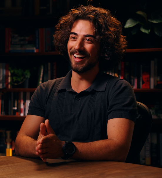 A smiling man with curly hair sits at a table in front of bookshelves during an interview.