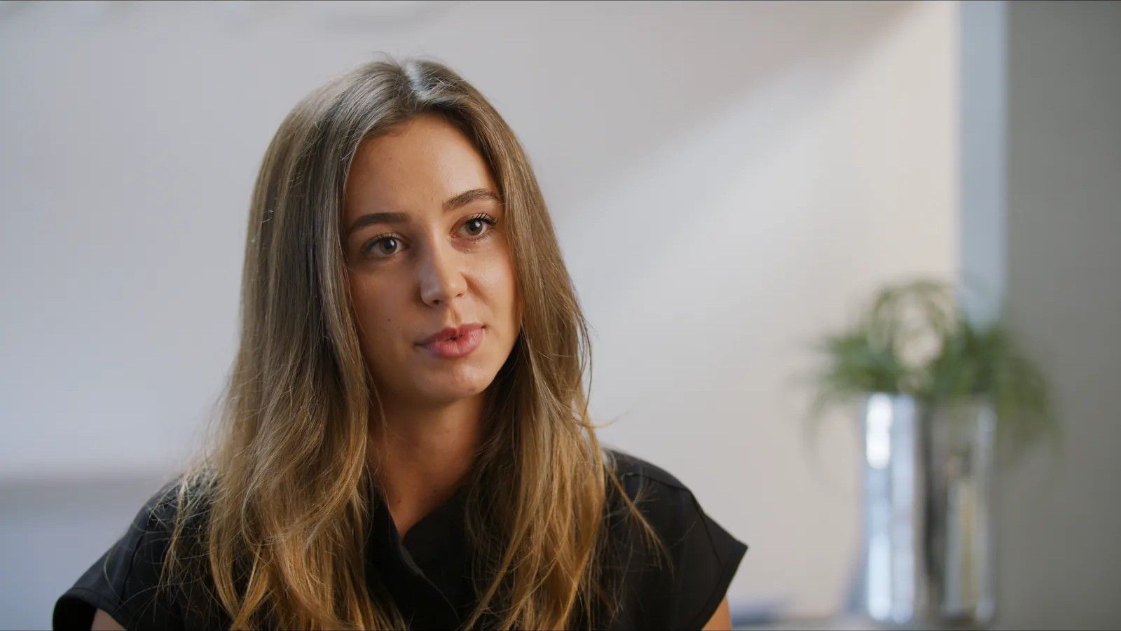 A woman with long hair speaks during an on-camera interview in a bright room.