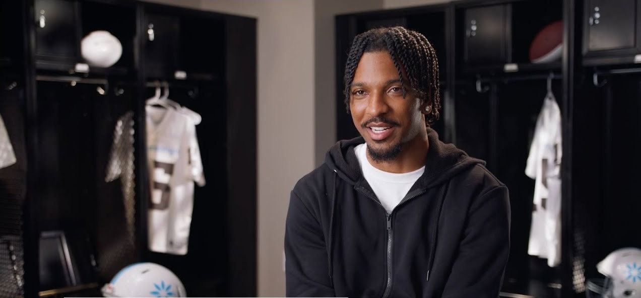 Jayden Daniels, in a black hoodie, smiles mid-interview in a locker room lined with white football jerseys.