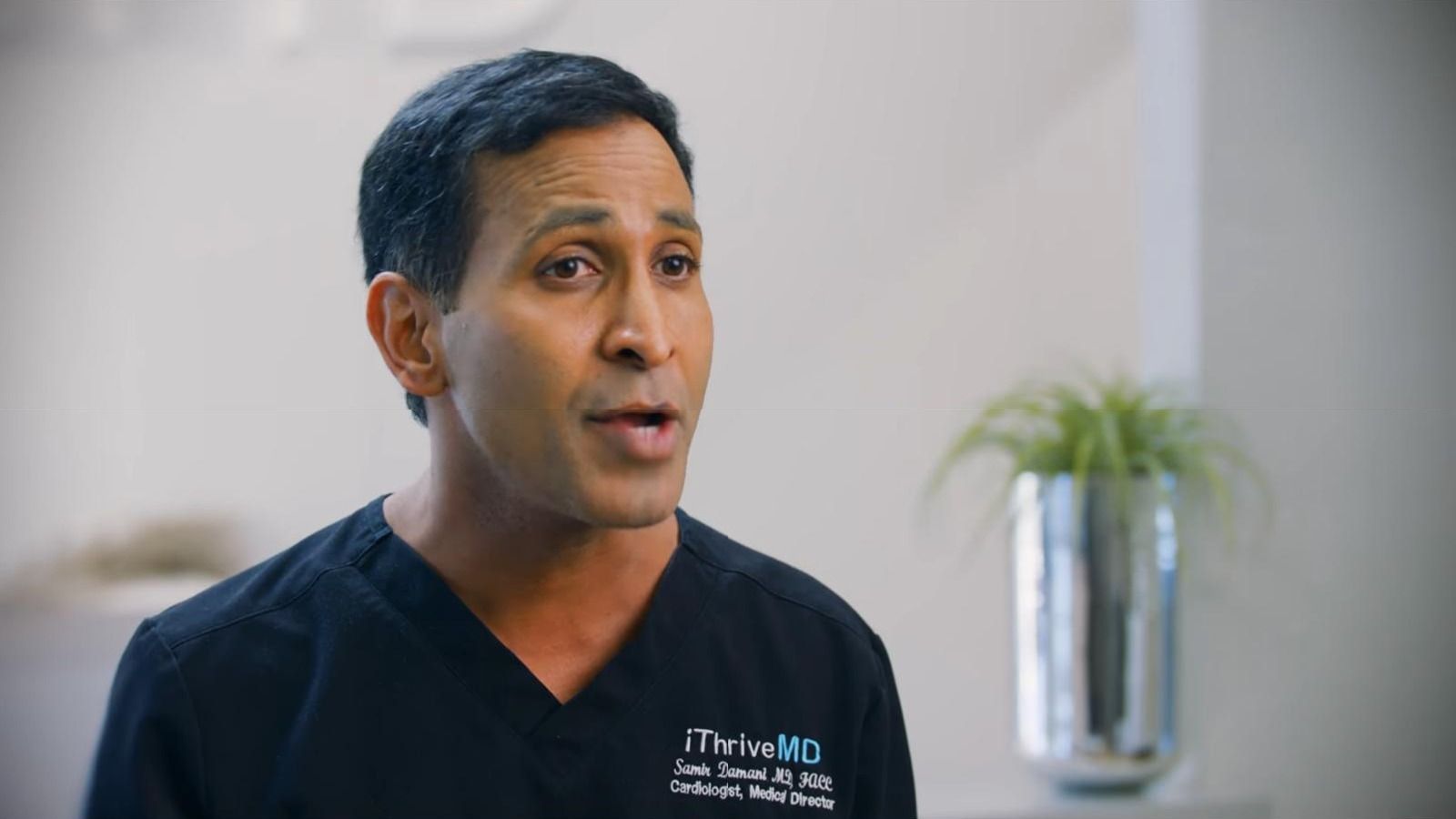 Dr. Samir Damani in navy iThriveMD scrubs speaks in an interview, a potted plant on a shelf behind.