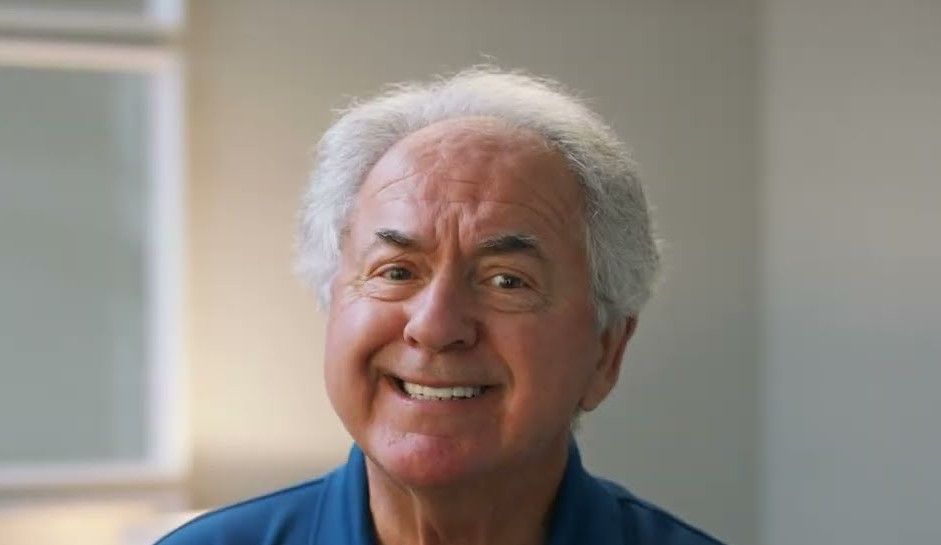 Wayne, a white-haired man in a blue polo, grins during his interview.