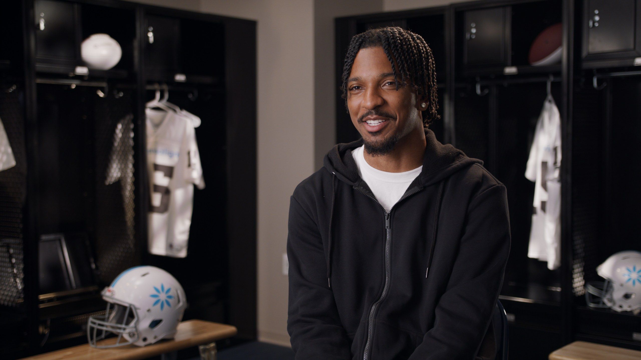 A smiling man in a black hoodie sits for an interview in a locker room, jerseys and helmets behind him