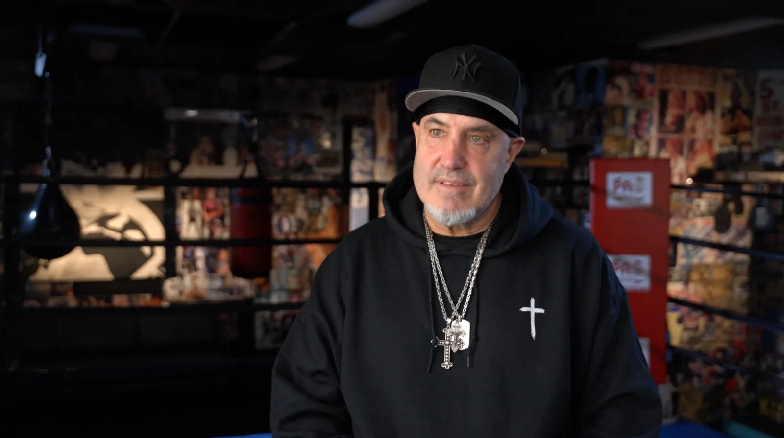 A man in a black cap and hoodie talks to camera in a boxing gym hung with fight posters.