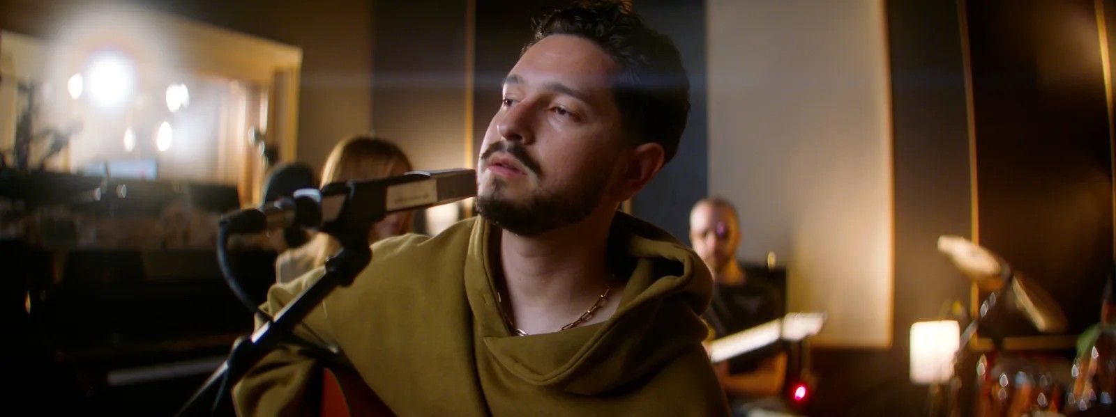 A singer in a hoodie plays guitar at a studio microphone, the band behind him in warm light.