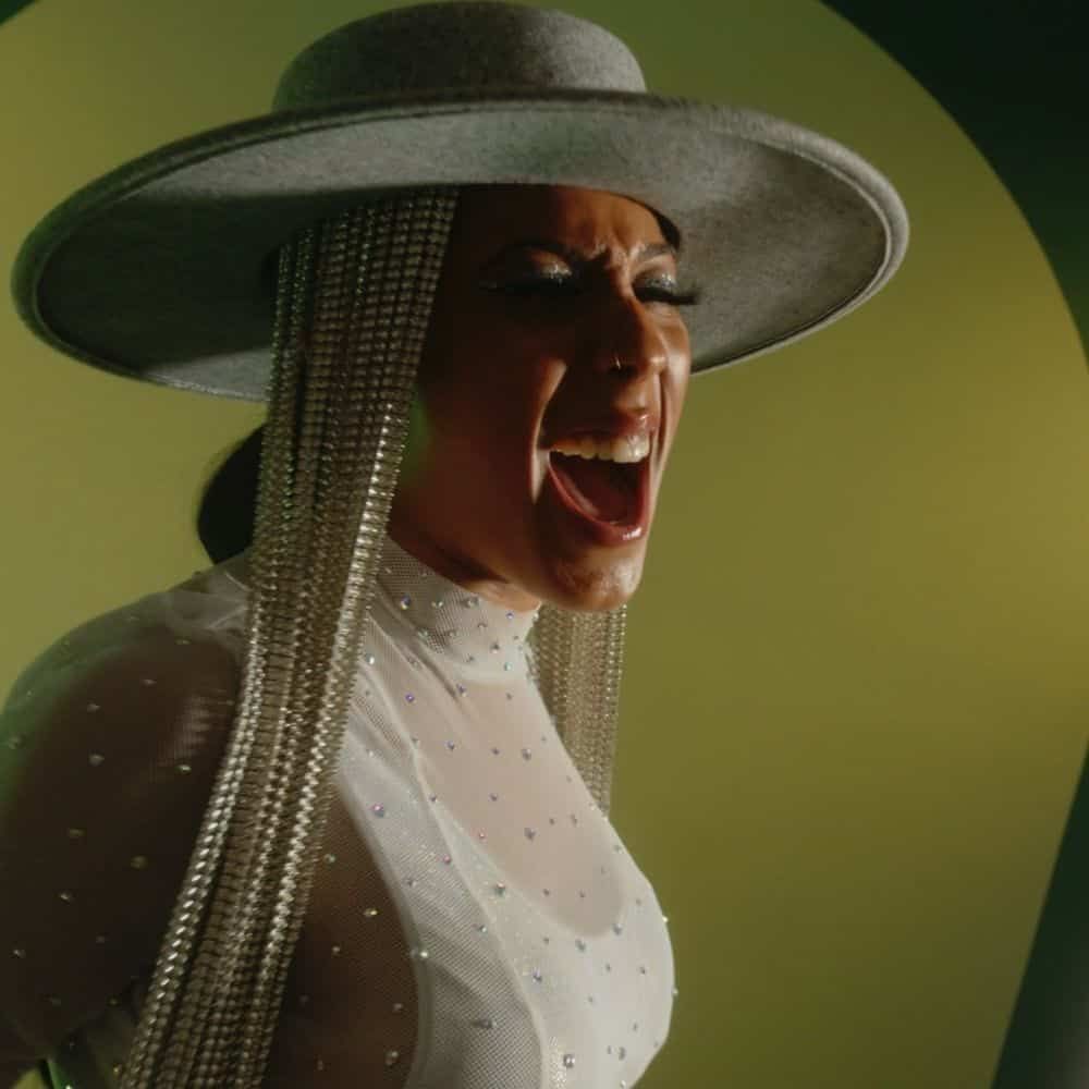 A singer in a wide-brimmed gray hat with long crystal fringe belts out a note against a green backdrop.