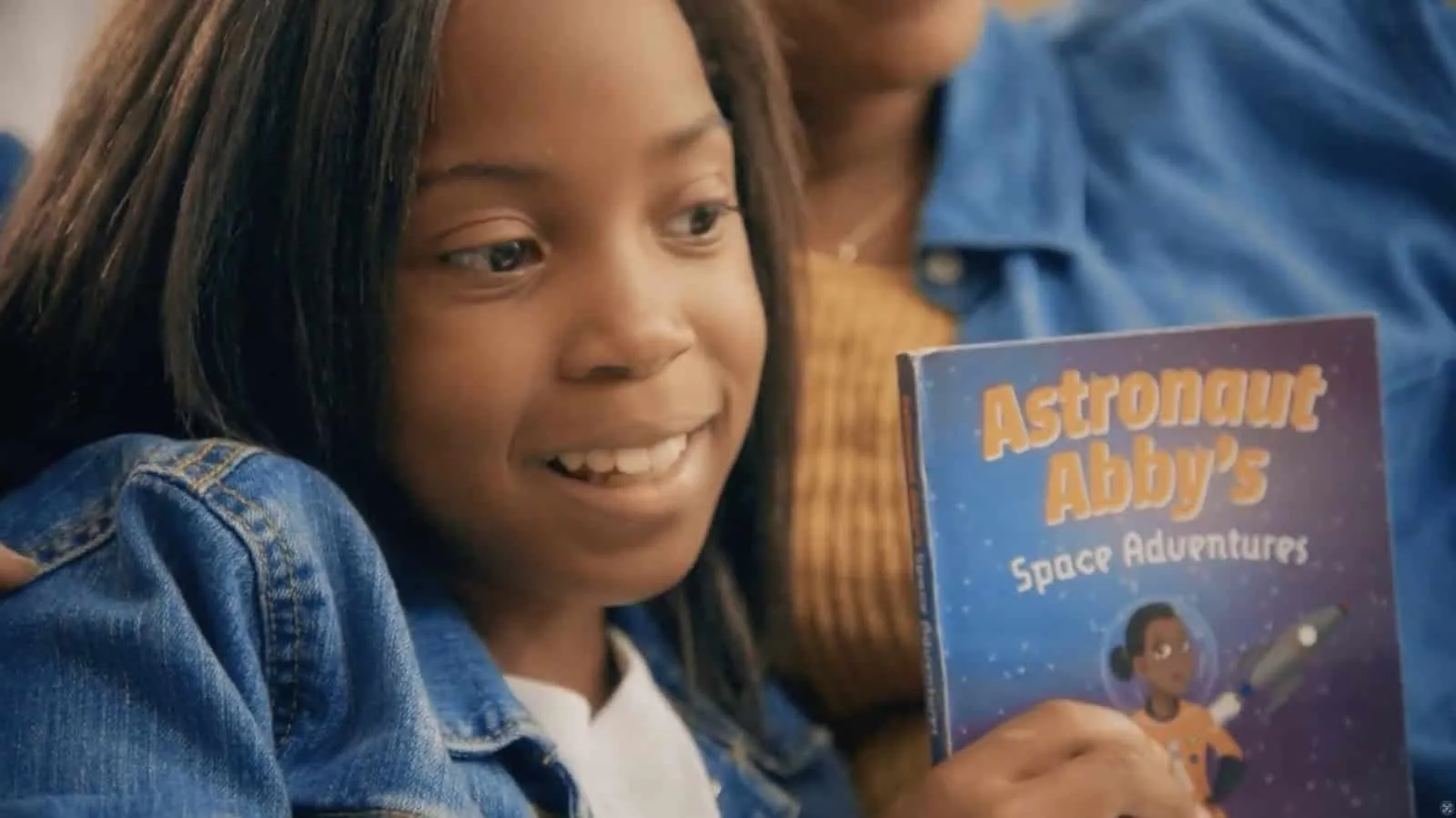 A smiling girl holds up the picture book Astronaut Abby’s Space Adventures.