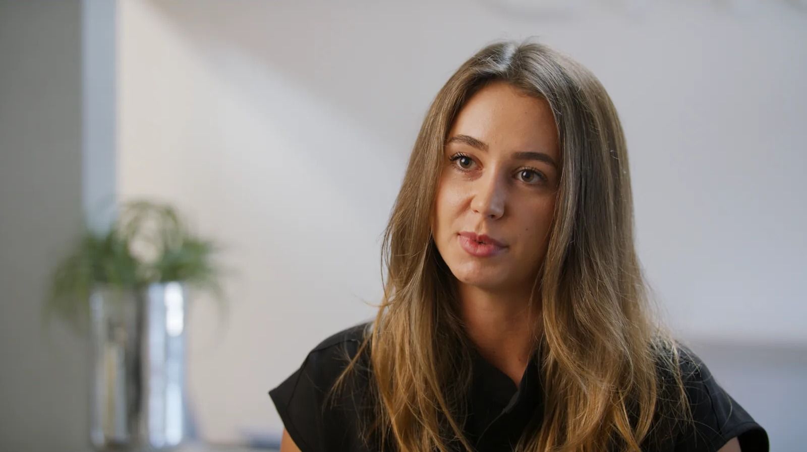 A woman with long brown hair tells her story on camera in a softly lit interview.