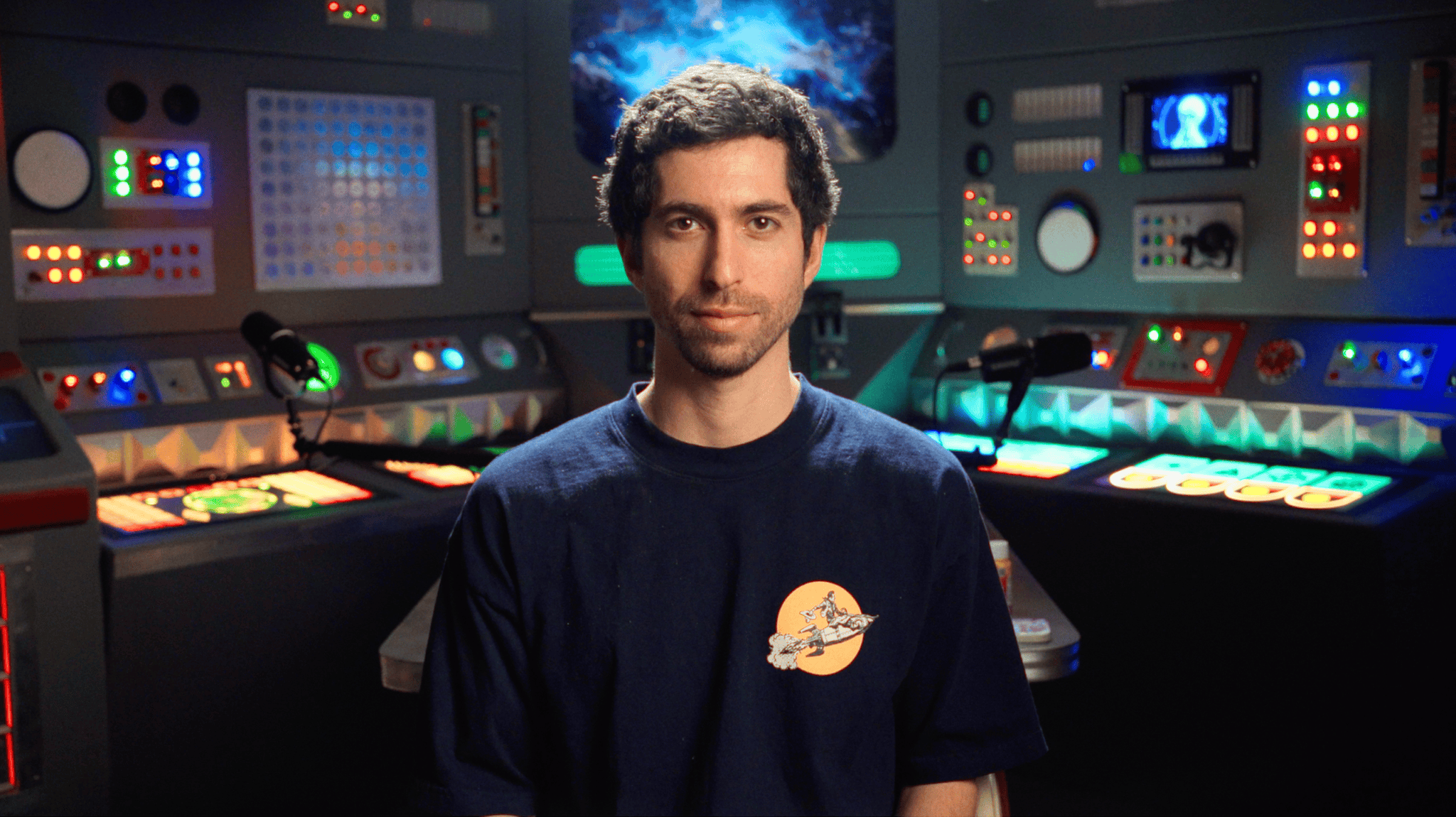 A host sits in front of a retro control-room set with glowing panels.