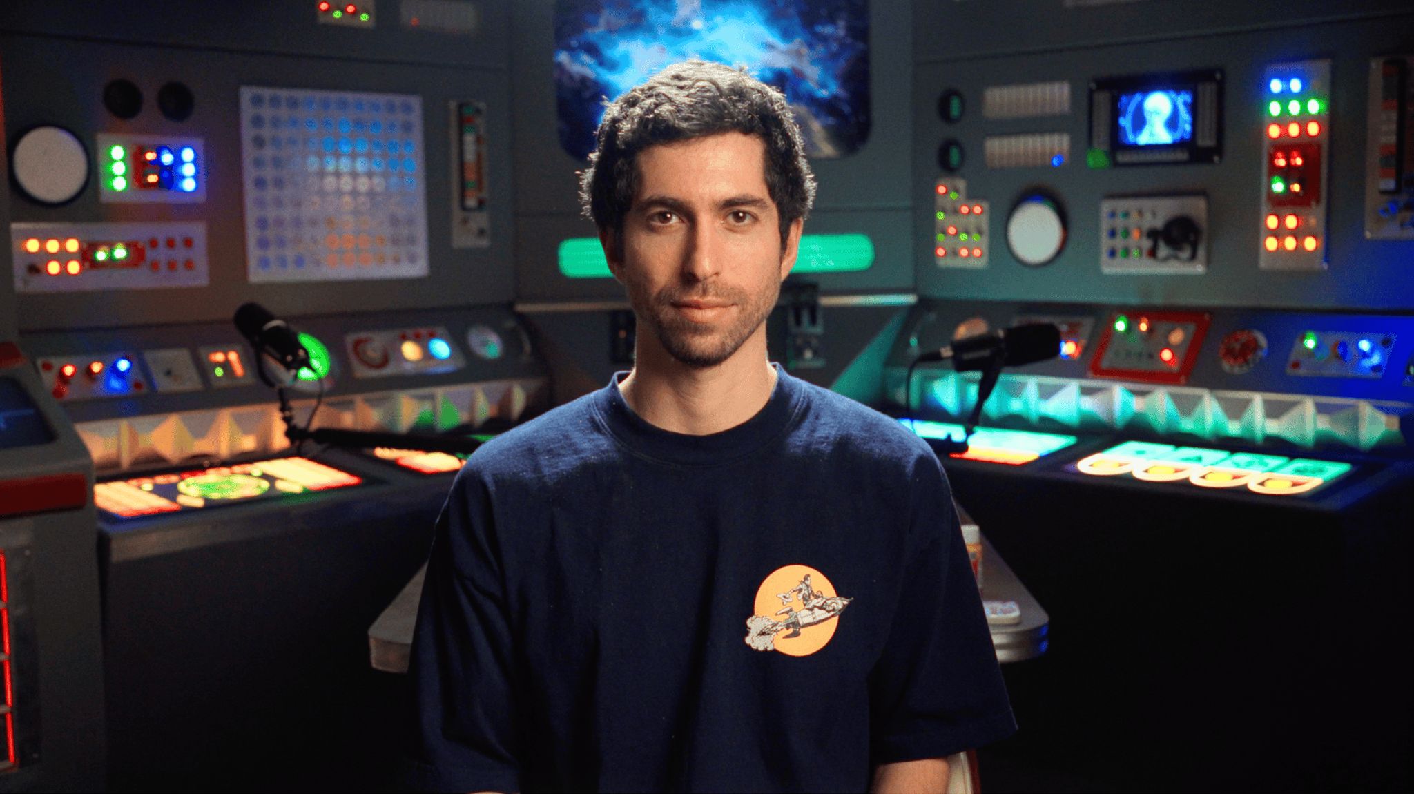 Jesse Michels sits in a retro-futurist podcast set lined with glowing control panels.