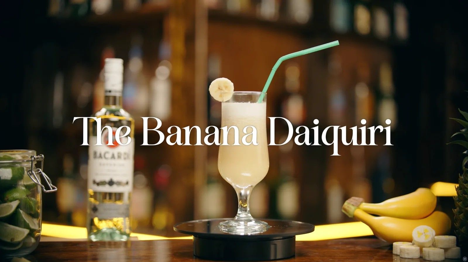A banana cocktail with a straw sits on a turntable at a bar beside a rum bottle, under the title "The Banana Daiquiri".