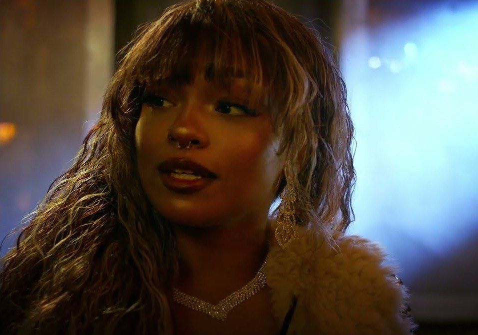 A model with feathered bangs, a nose ring and a fur collar looks off camera in warm light against a blue background.