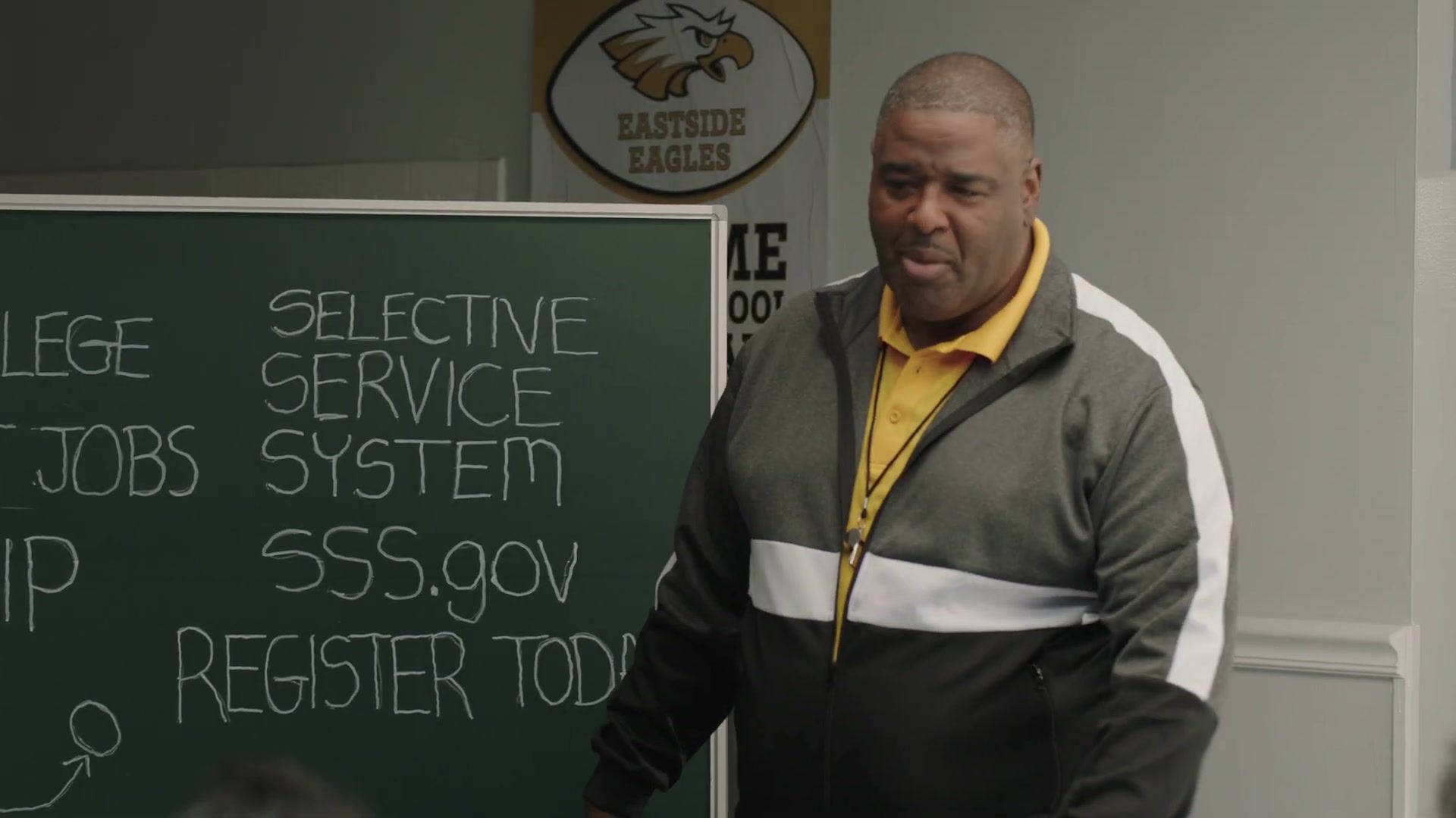 A coach in a track jacket stands at a chalkboard that reads Selective Service System, SSS.gov, Register today.
