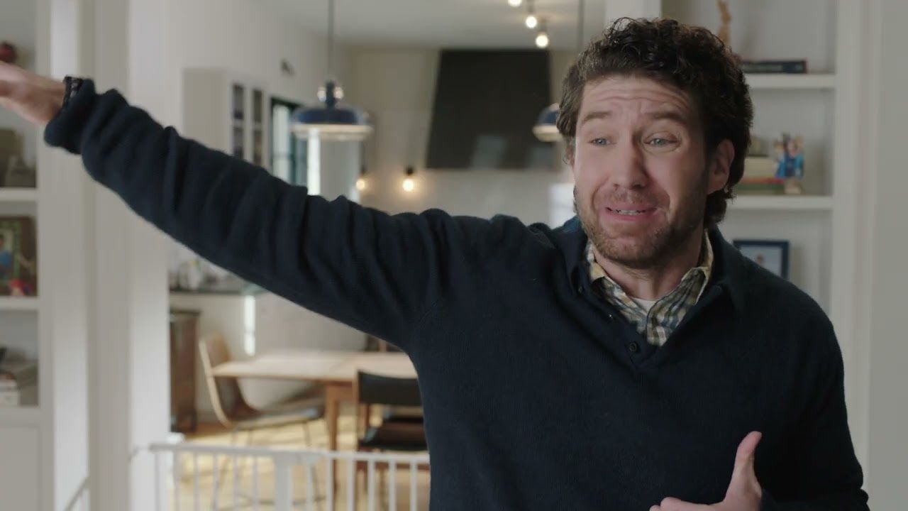 A man in a navy sweater flings one arm out wide and grimaces mid-sentence in a bright family kitchen