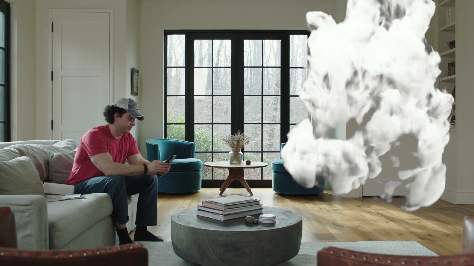 A man in a red T-shirt and cap sits on a sofa as a cloud of smoke bursts into his living room.