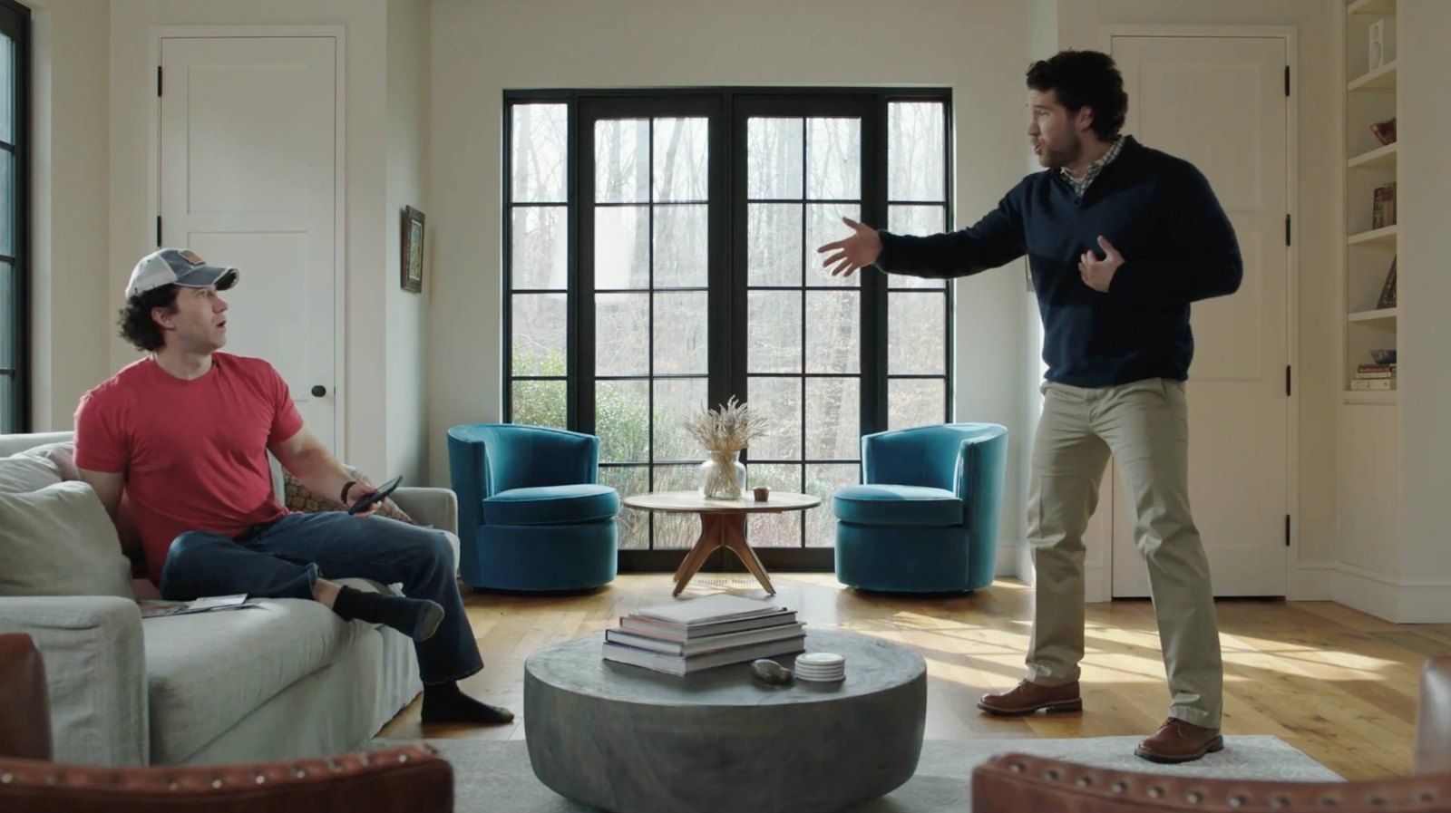 In a bright living room, a man in a navy sweater talks with his hands to a man in a red T-shirt and cap on the sofa.