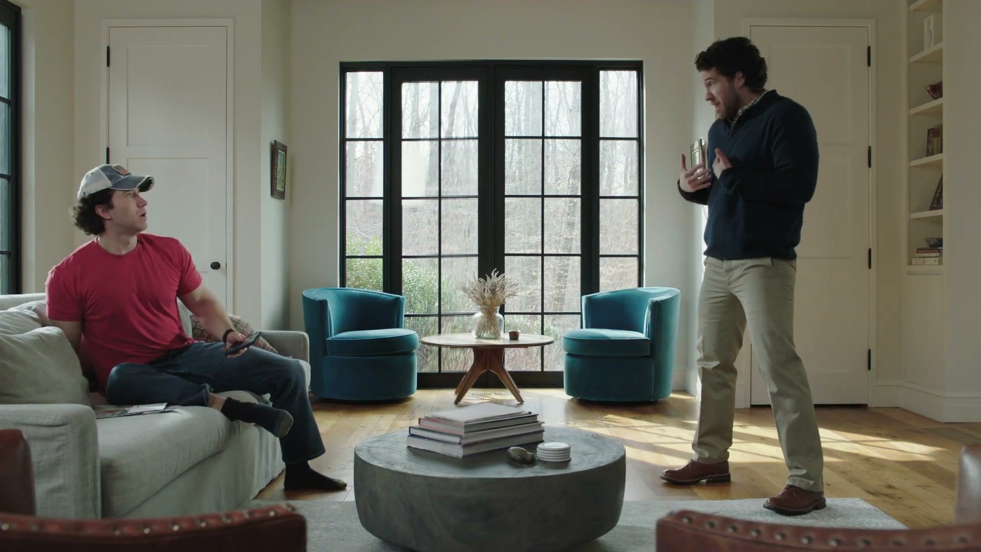 In a sunlit living room, a man in a red T-shirt and cap sits on the sofa while another man stands and talks with his hands.